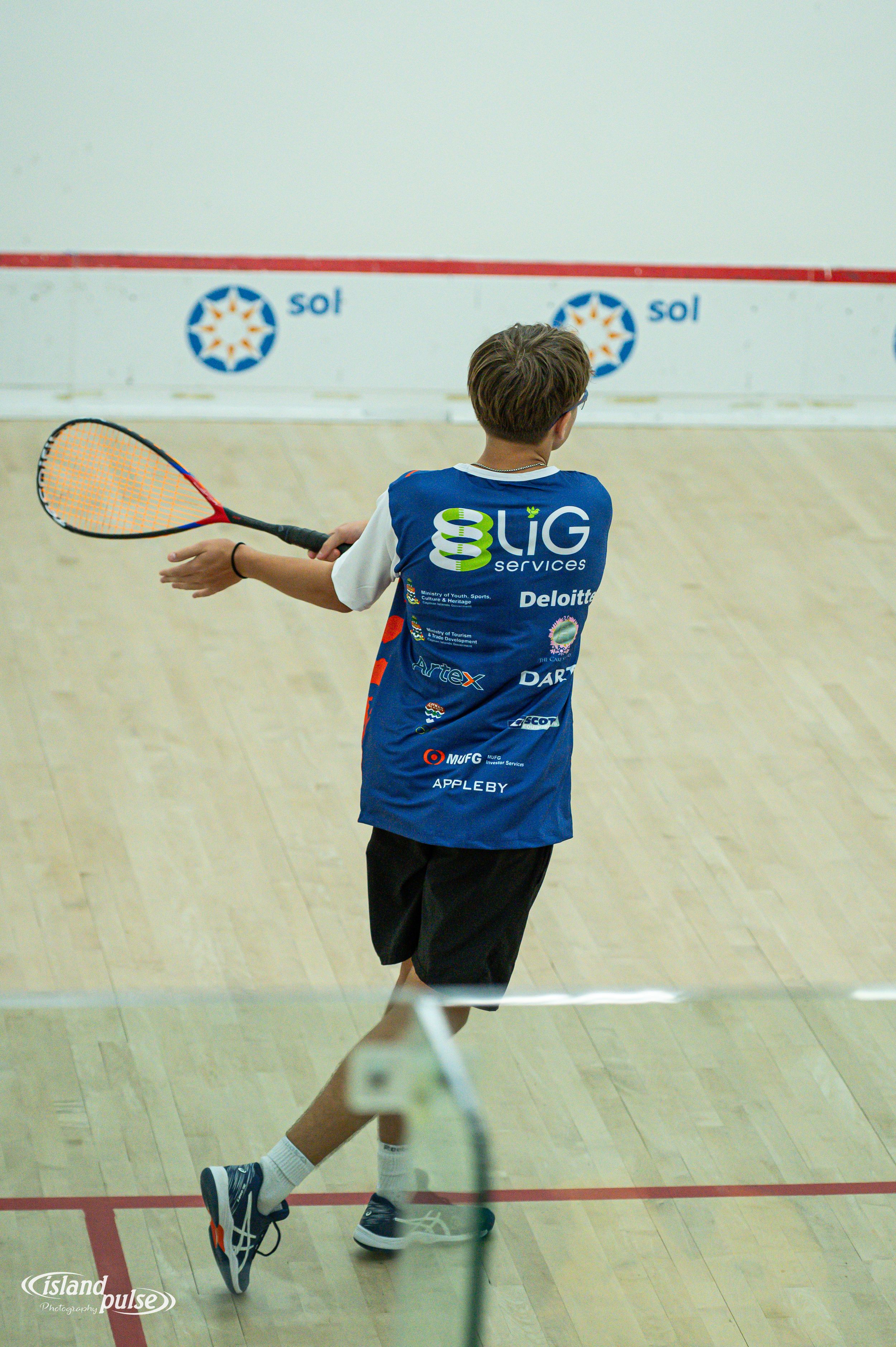 Young squash player in blue BUG Services jersey mid-swing on indoor court during competitive match.