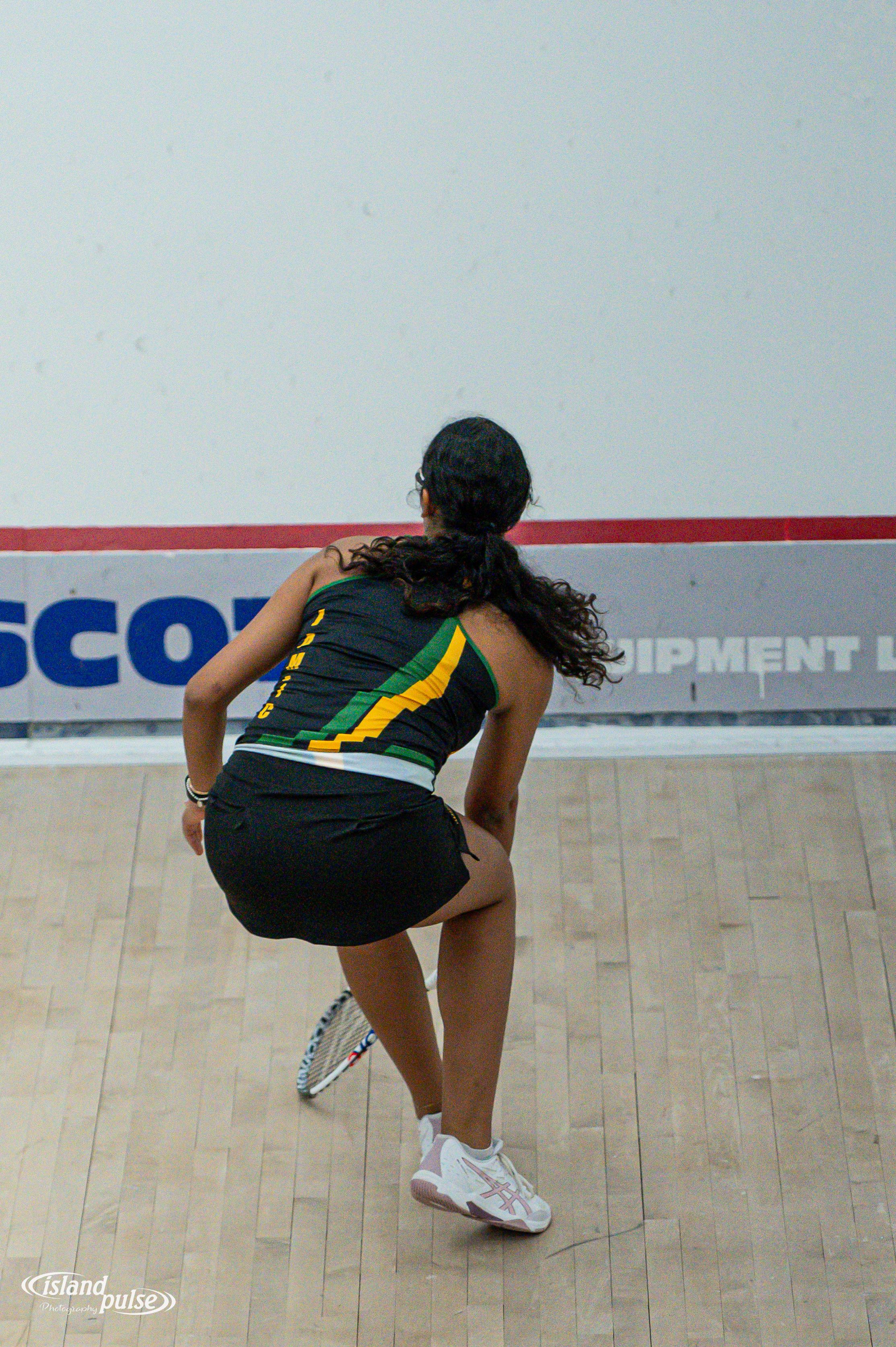 Female squash player in green and black jersey crouches in ready position on indoor court during competitive match.