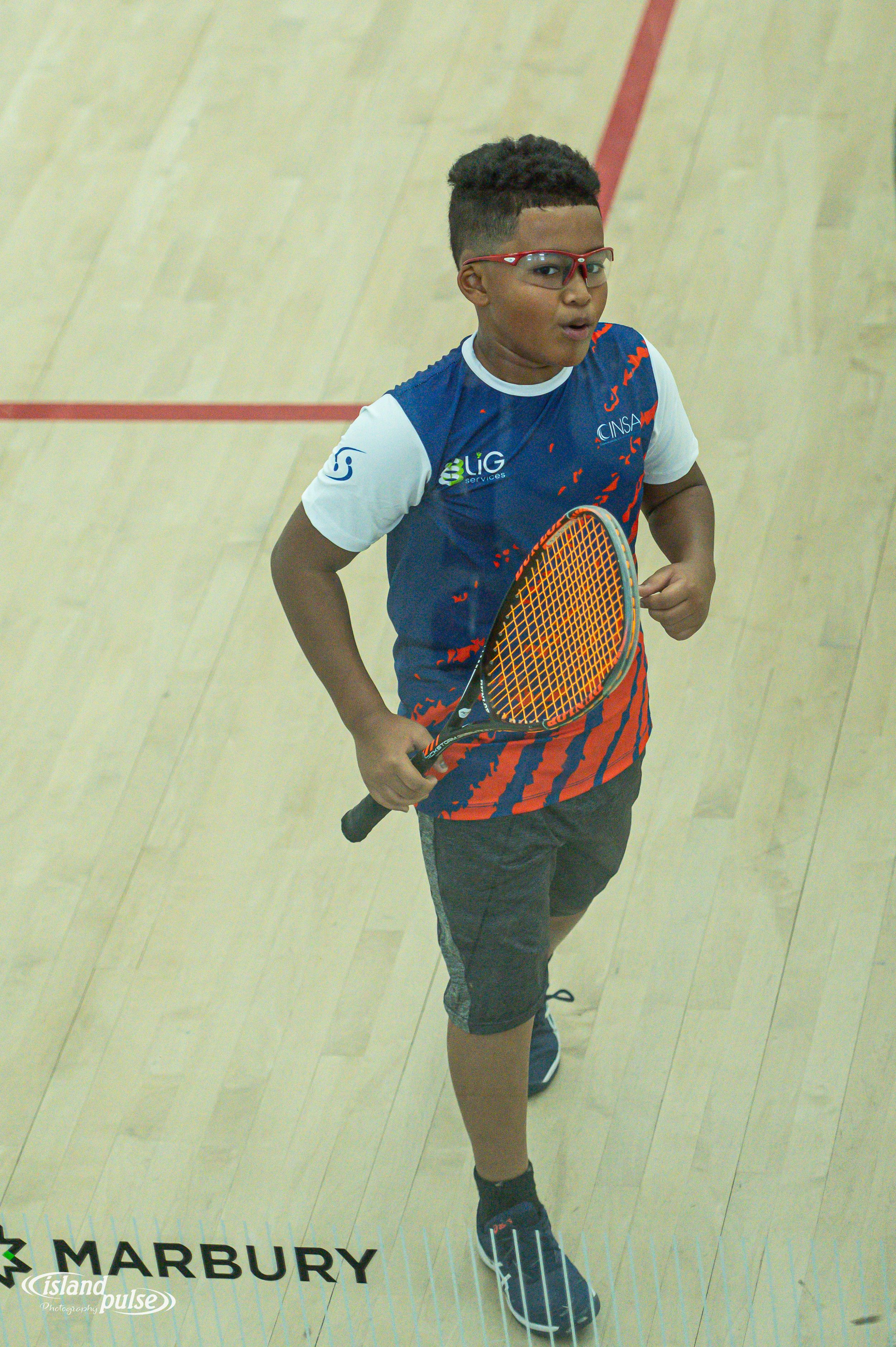 Young boy in blue sports jersey playing squash on indoor court, holding racket and wearing red glasses.