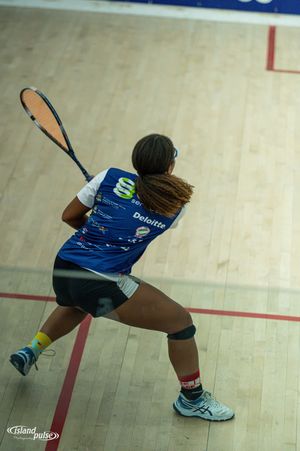 Female squash player in blue Deloitte jersey mid-swing on indoor court, viewed from behind.