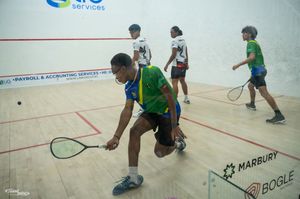 Players competing in an indoor squash court match, with one athlete lunging low to return the ball.