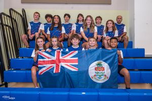Youth sports team posing with Cayman Islands flag in blue bleachers wearing matching uniforms.