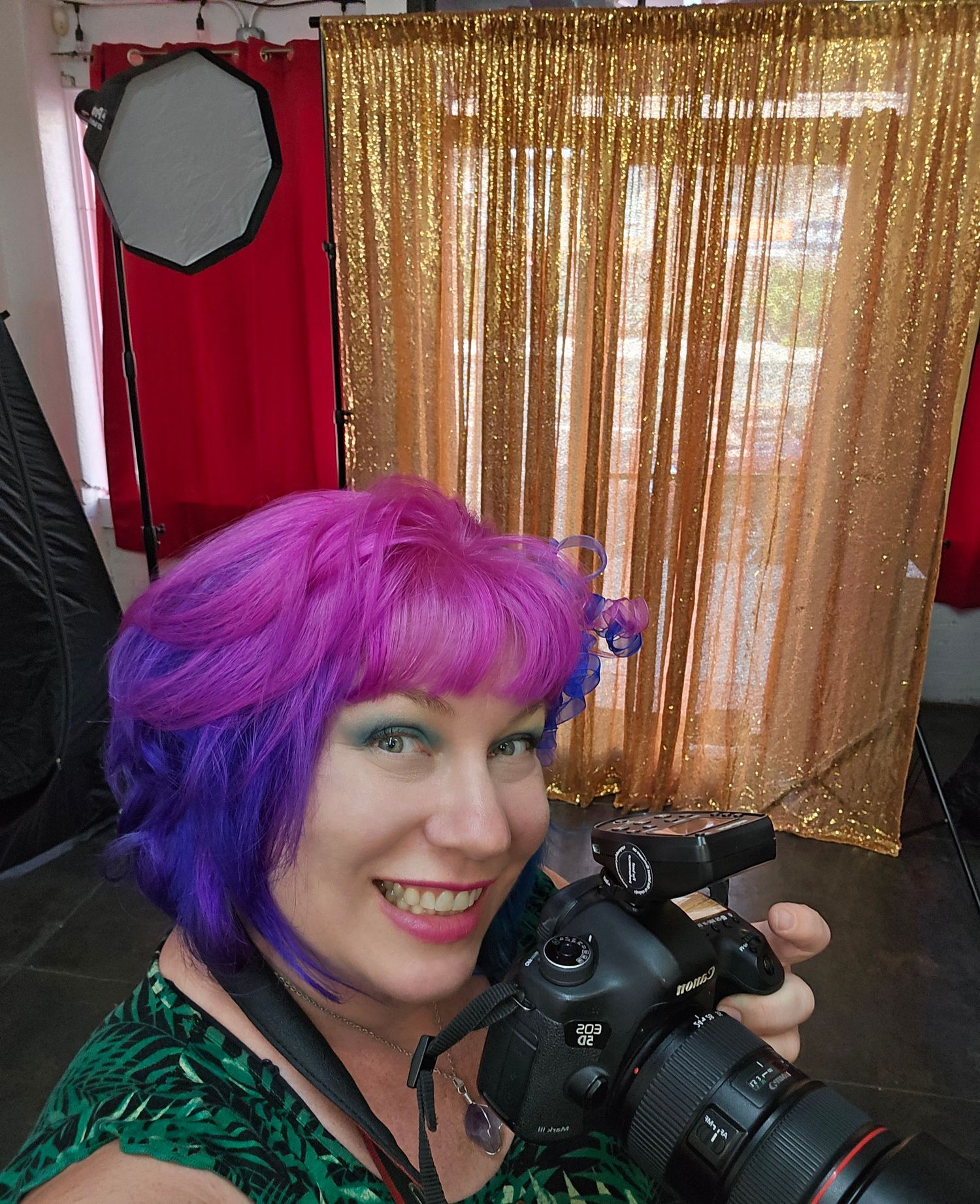 Photographer with bright purple and pink hair holding camera against sparkly gold backdrop.