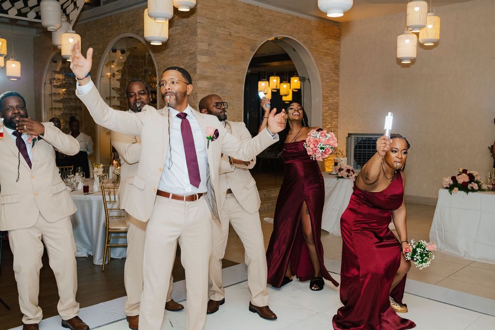 Wedding guests dancing joyfully at a reception under elegant pendant lighting in an arched venue.