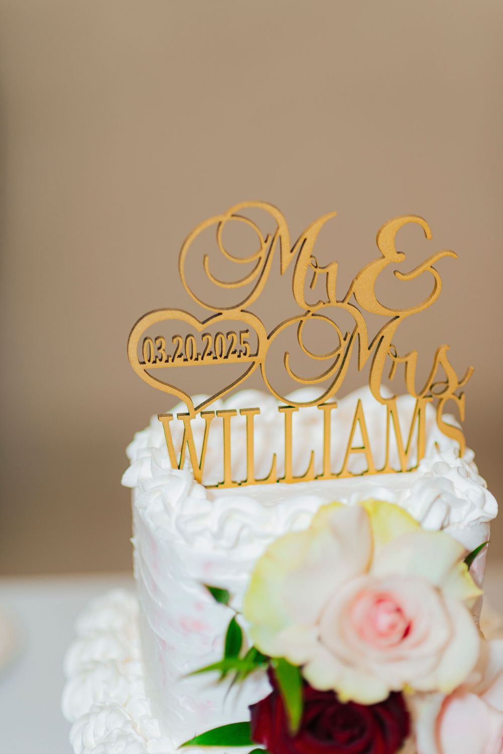Gold wedding cake topper with 'Mr & Mrs Williams' text sits atop white frosted cake decorated with pink and red roses.