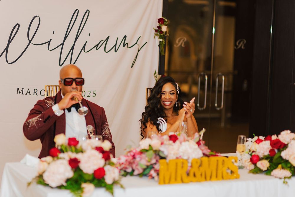 A couple sits at an elegant wedding reception head table decorated with pink and white floral arrangements.