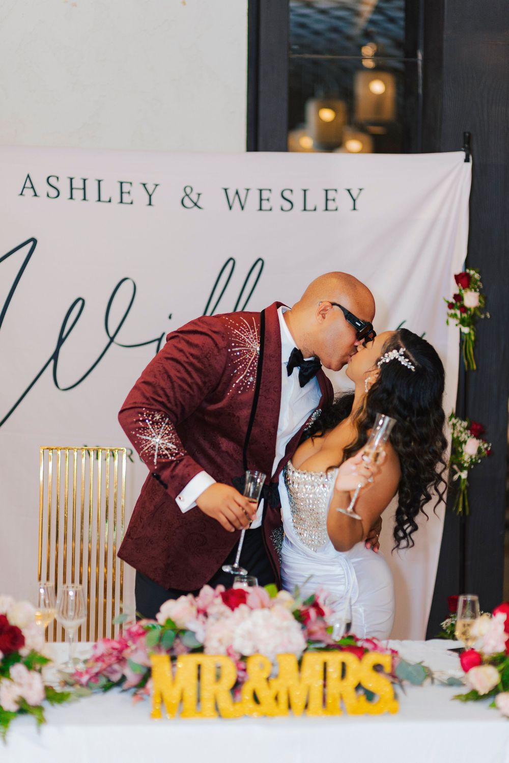 Wedding couple shares a romantic kiss at their reception in front of a personalized banner and floral decorations.