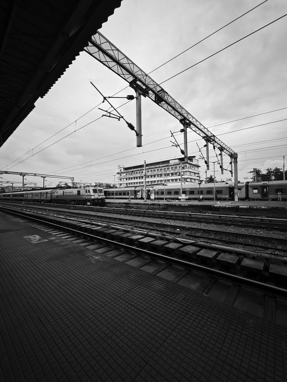 Black and white view of a railway platform with overhead power lines and a multi-story building in the background.