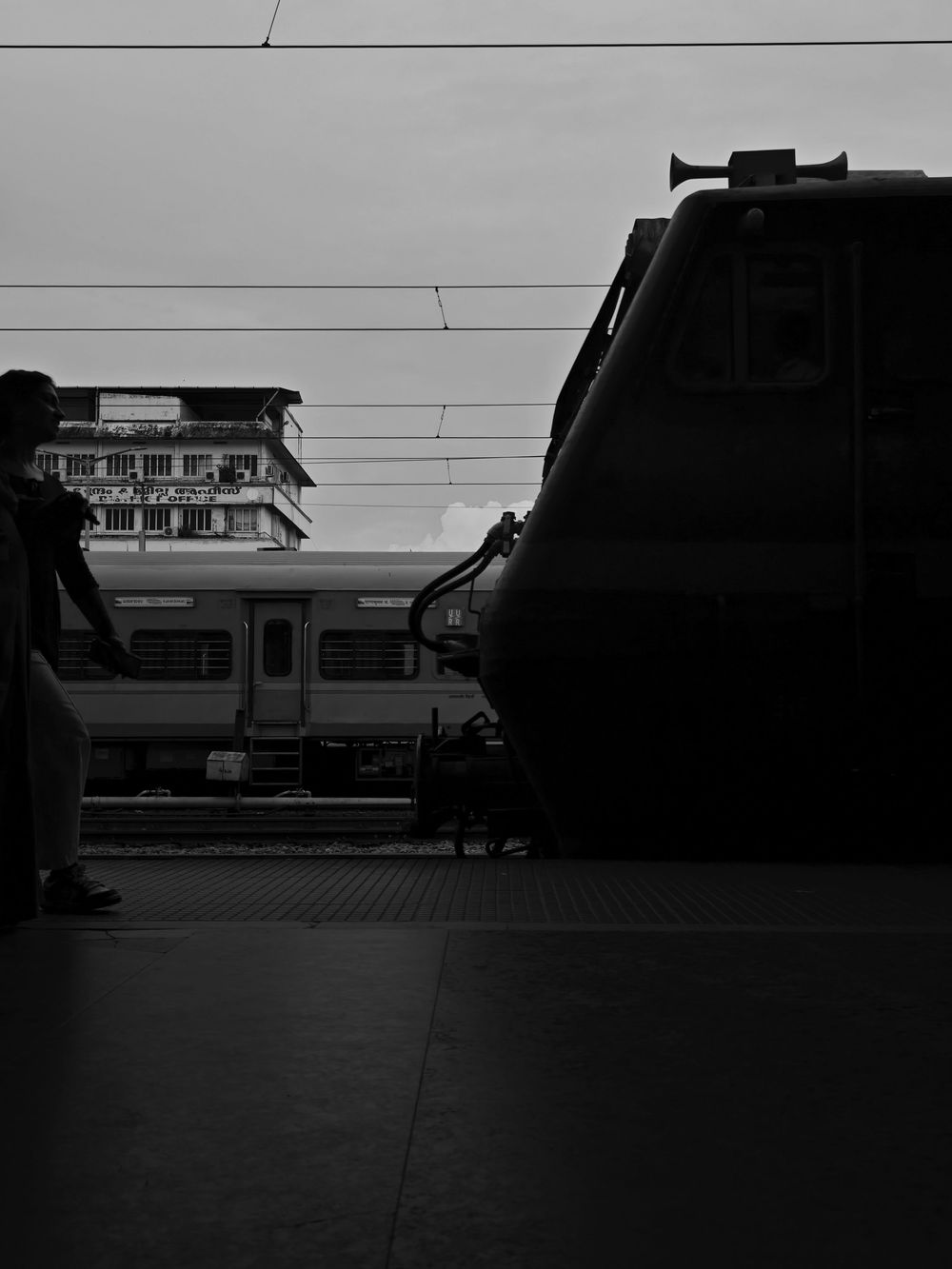 A silhouetted train platform with overhead power lines and a commuter train visible in black and white photography.