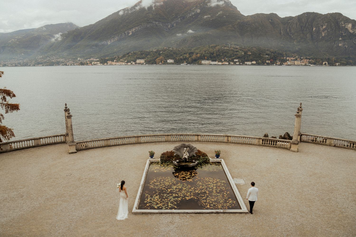 Scenic lakeside terrace with ornate stone railings and garden feature overlooking mountain landscape.