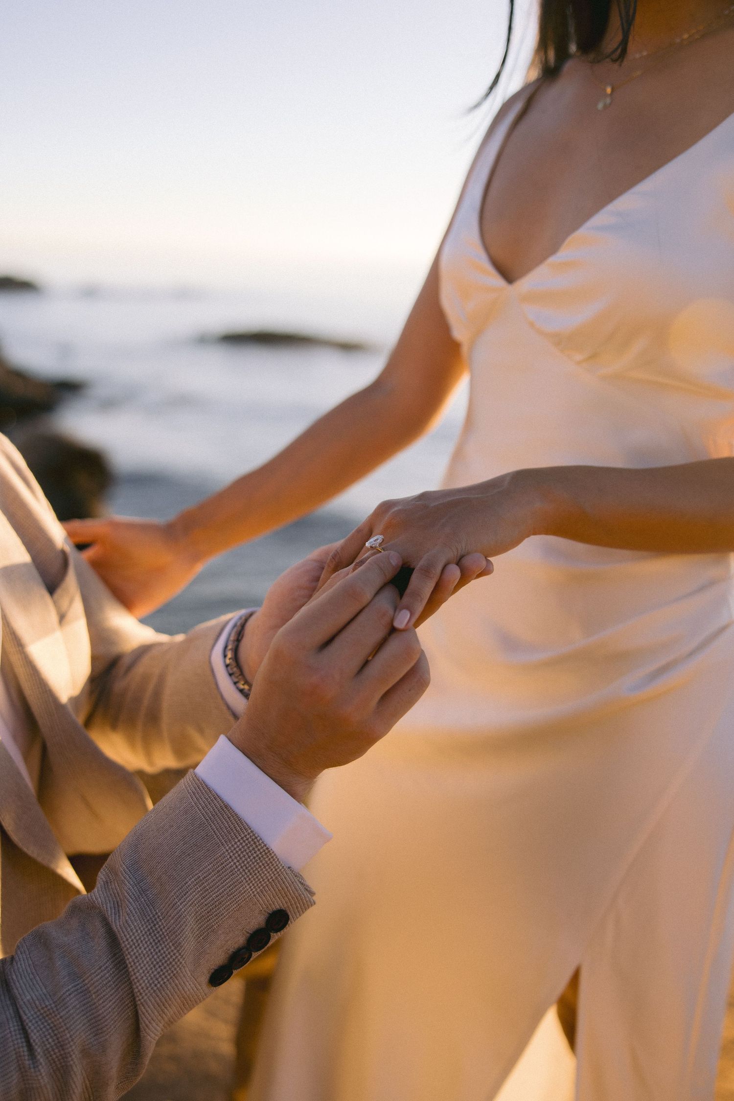 A close-up shot of hands holding during a beach wedding ceremony at sunset, with the bride wearing a white dress.