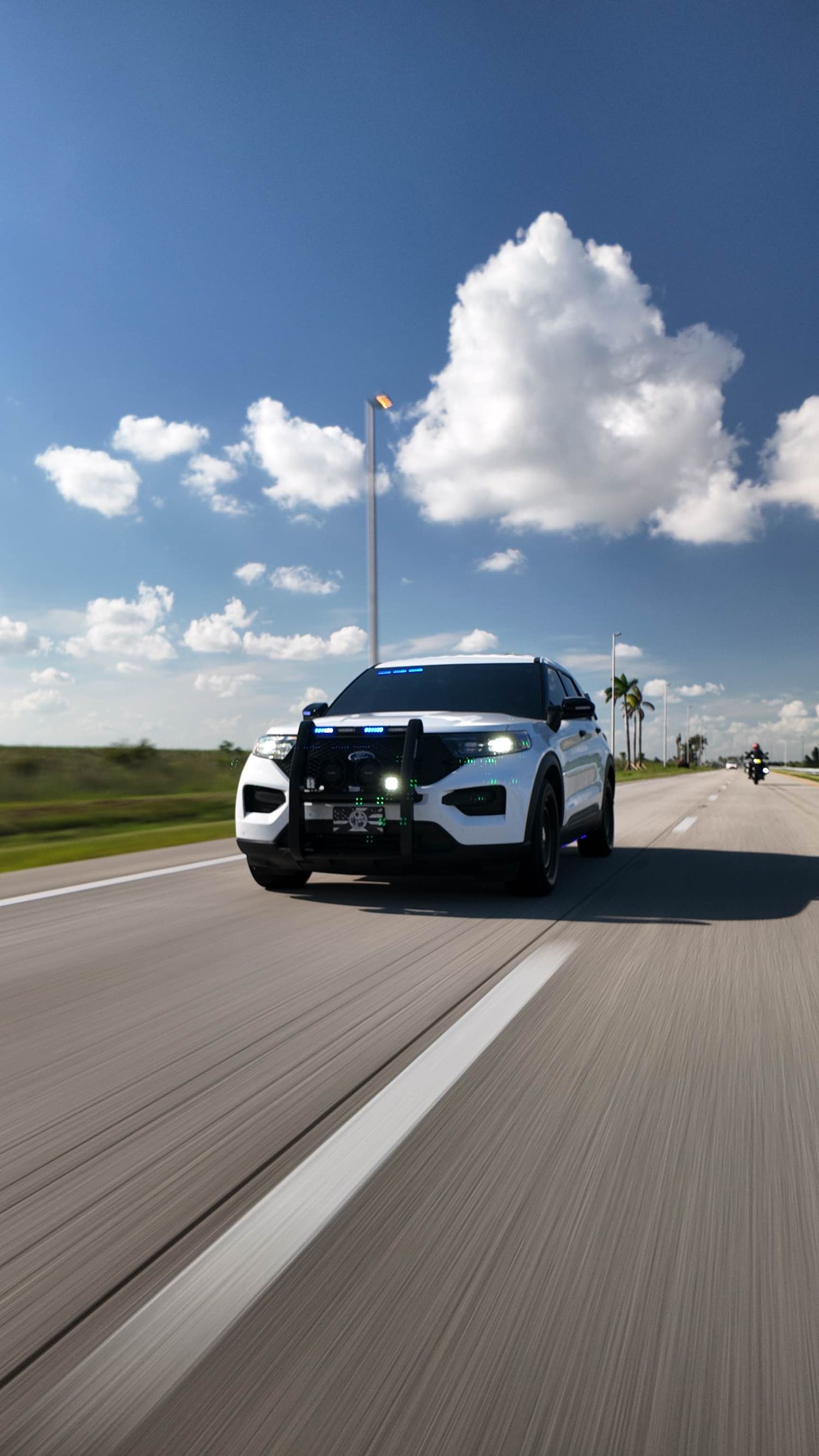 A white police interceptor vehicle drives on a road under puffy clouds and blue sky.