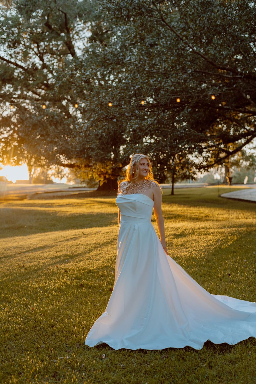 Bride in flowing white gown poses in golden sunset light under a large tree.