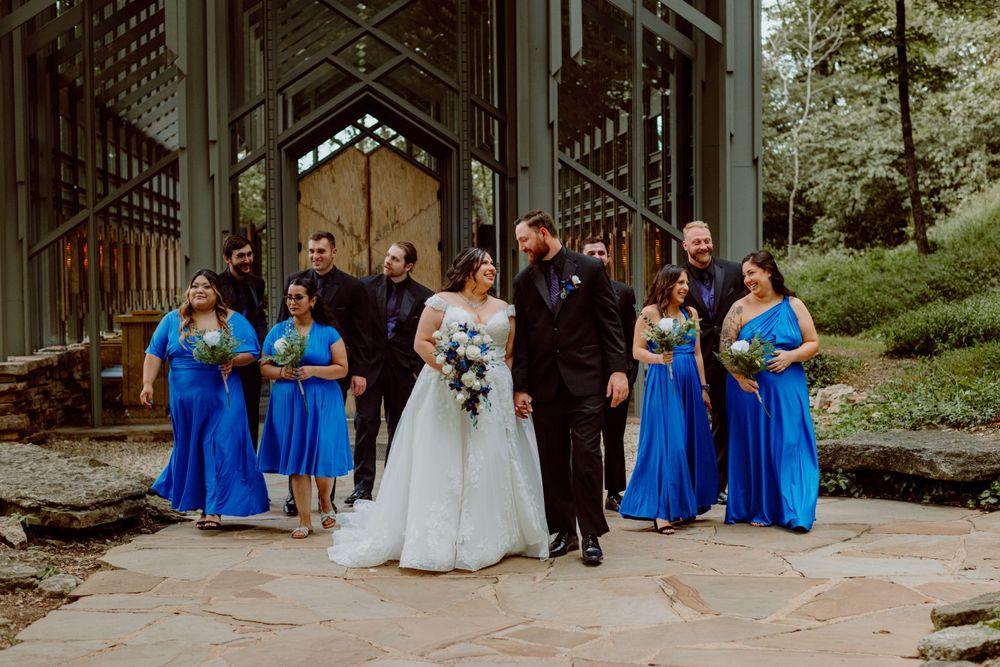 Wedding party poses in formal attire with bridesmaids in blue dresses and groomsmen in black suits.