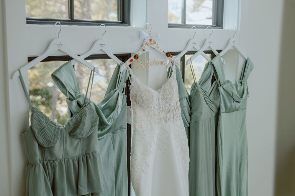 Elegant wedding dresses hanging by a window with sage green bridesmaid gowns and a white lace bridal gown in the center.