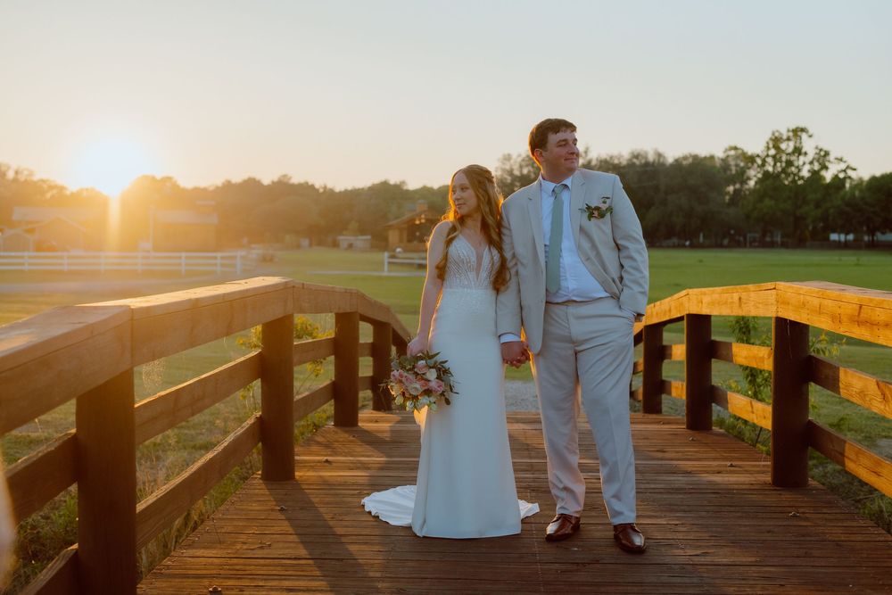 A couple in wedding attire stands on a wooden bridge during golden hour sunset in a scenic outdoor setting.