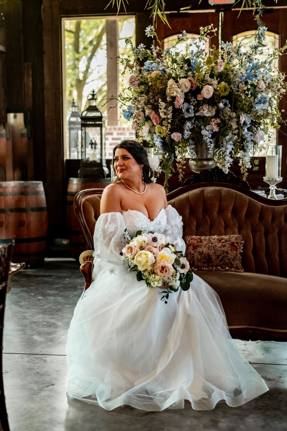 Bride sitting on vintage couch surrounded by elegant floral arrangements.