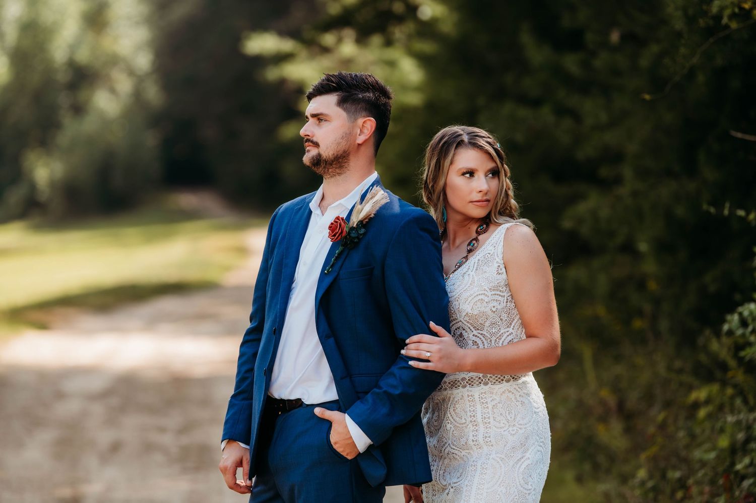 A couple in wedding attire poses together on a countryside path.