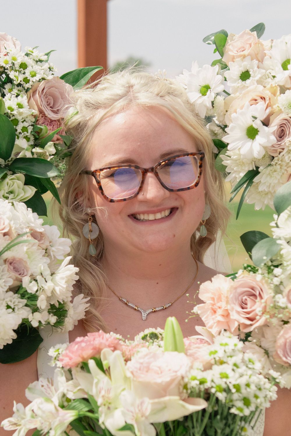 Smiling bride with tortoiseshell glasses surrounded by blush pink and white floral arrangements at outdoor wedding ceremony.