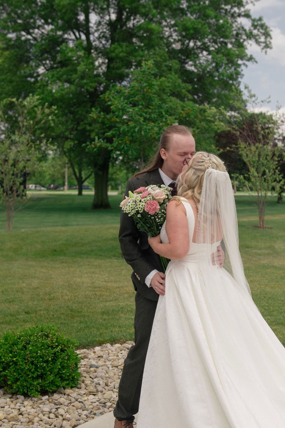 Bride and groom share a kiss outdoors on their wedding day, surrounded by lush green trees and manicured lawn.