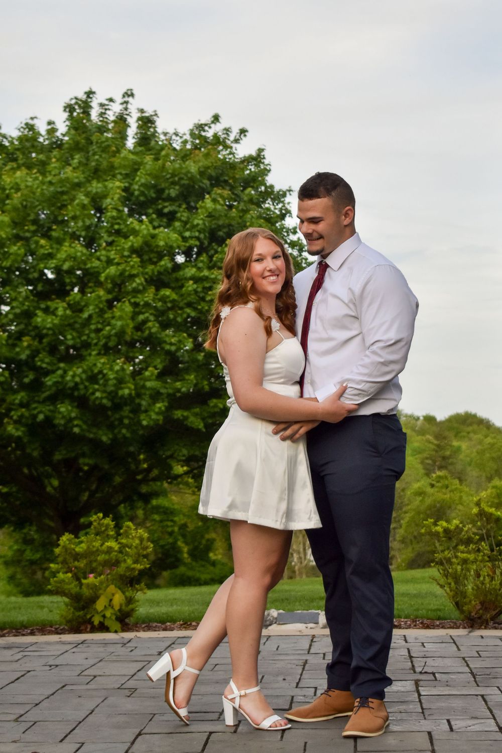 A couple shares a romantic moment on a stone patio with lush green trees in the background.