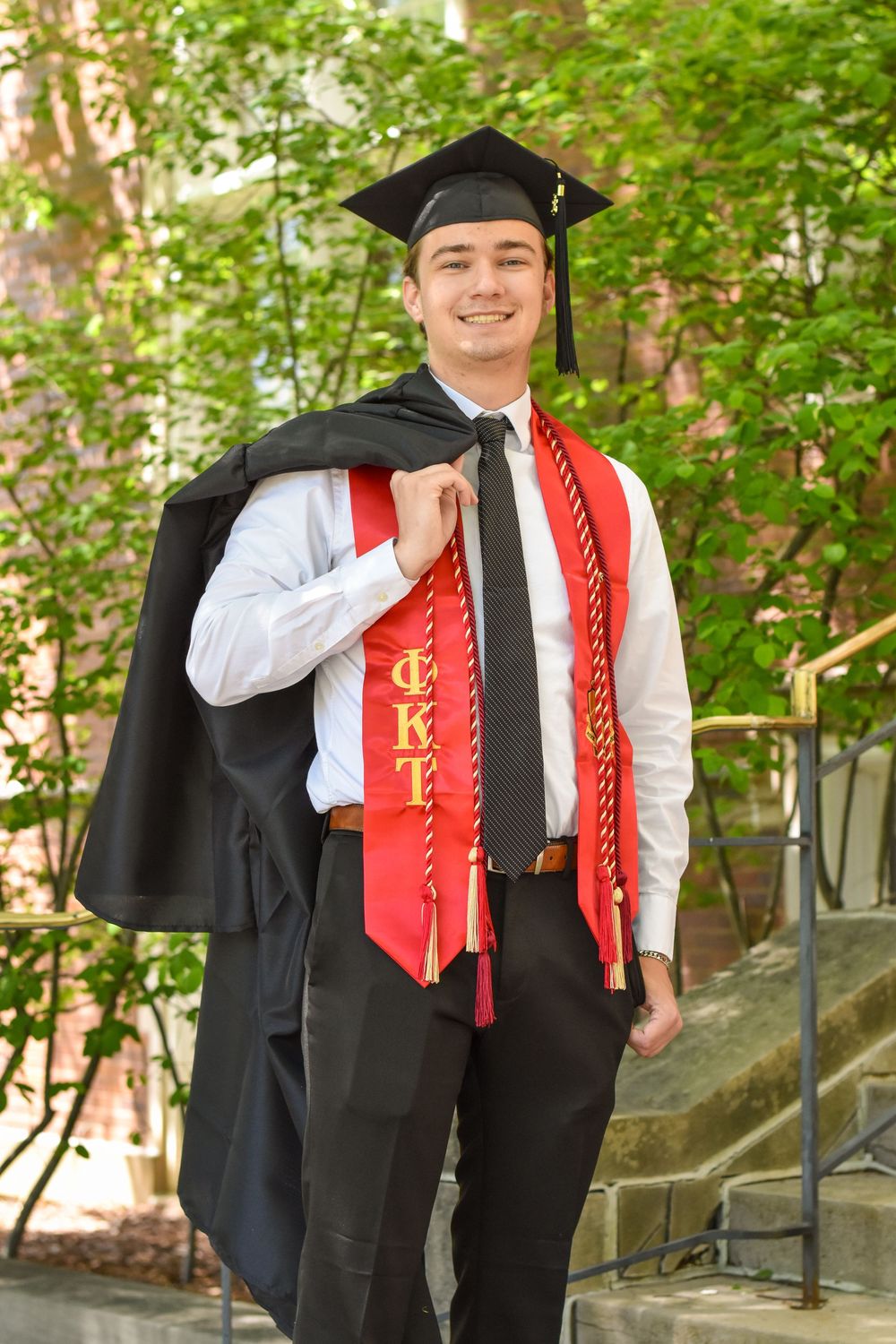 Graduate student in cap and gown with red honor cords poses outdoors among trees and greenery.