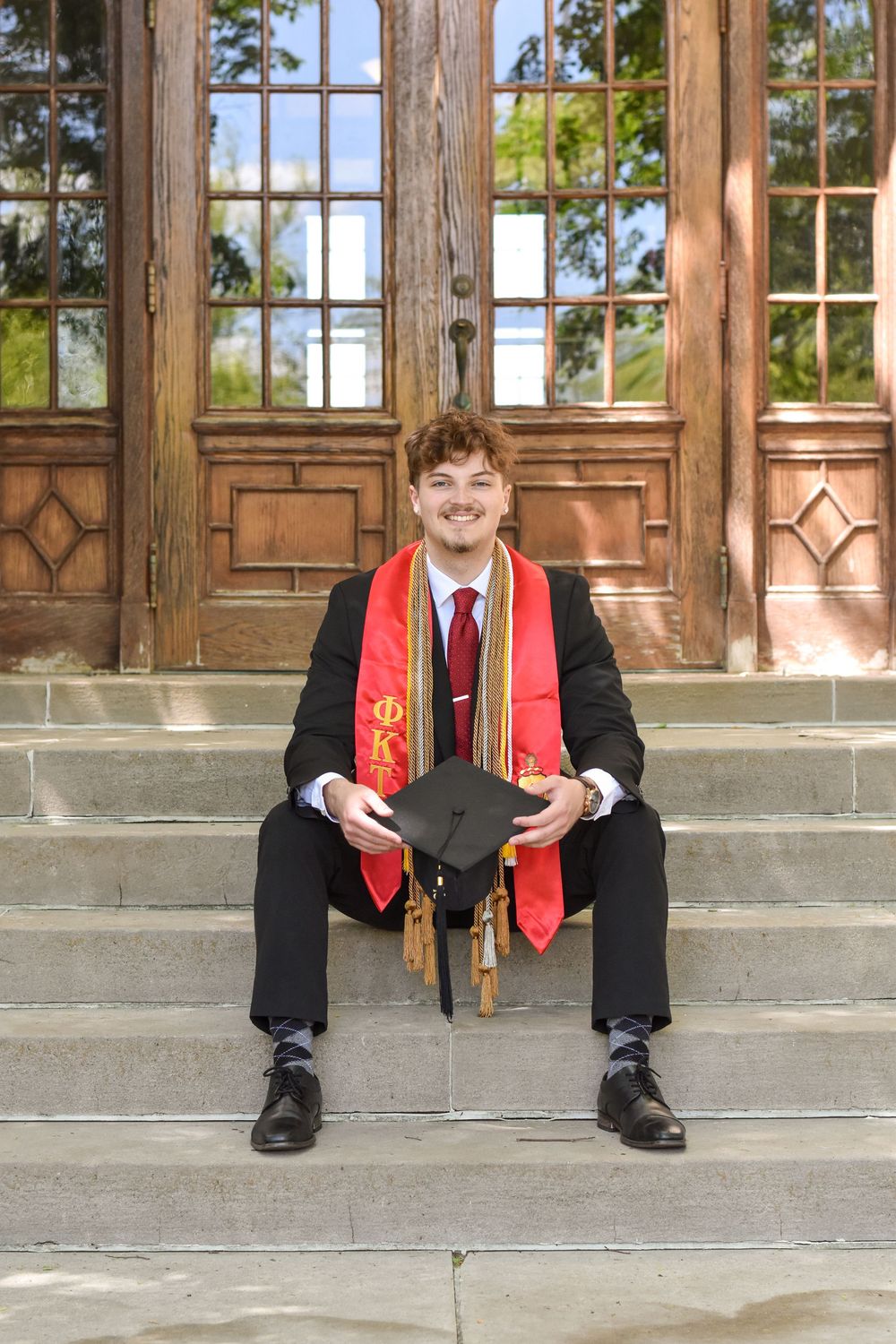 Graduate in academic regalia sits on stone steps in front of ornate wooden doors.