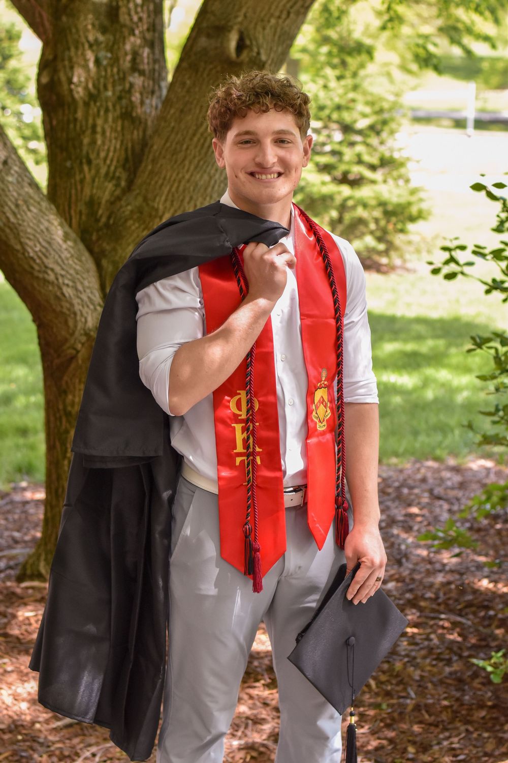 A graduate in black graduation gown and red honors stole stands smiling under a tree while holding their mortarboard.