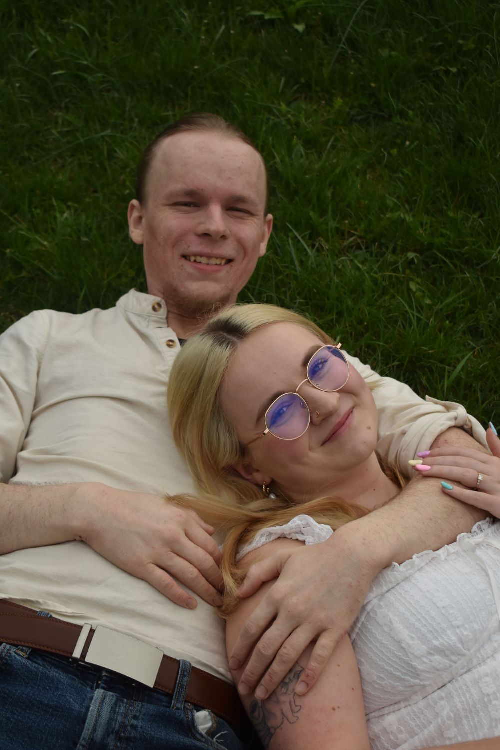 A couple relaxes together on green grass, with one wearing glasses and a white top while the other wears a light colored shirt.