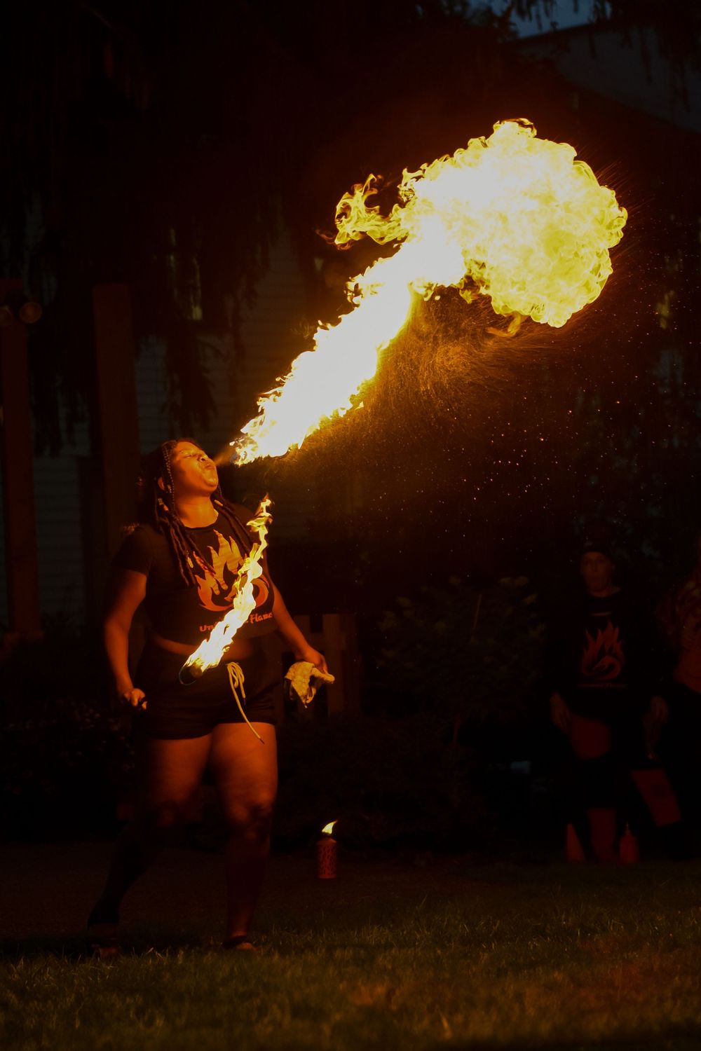 A fire breather performs a dramatic flame stunt at night, exhaling a large burst of fire into the dark sky.