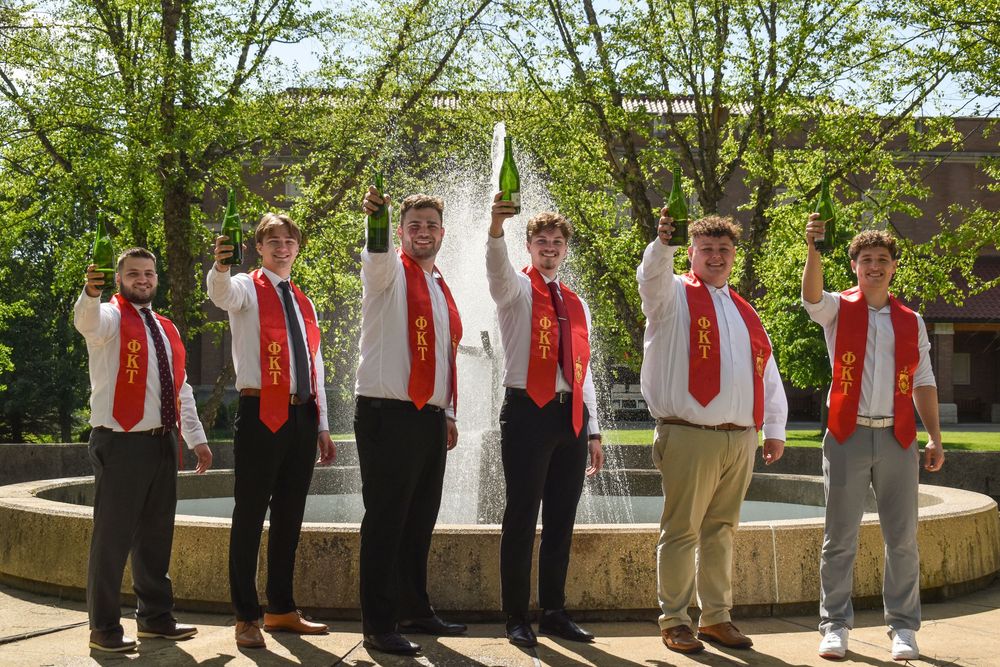 A group stands in formal attire with alternating red and white dress shirts against an outdoor backdrop.
