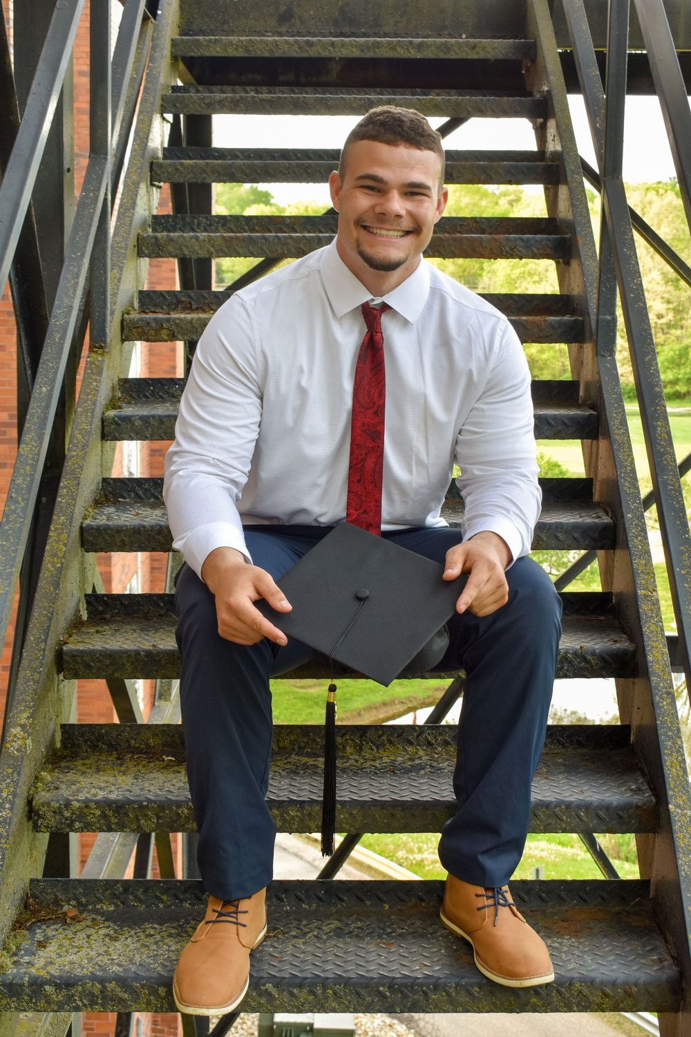 Graduate sitting on metal stairs holding mortarboard cap wearing white dress shirt and red tie.