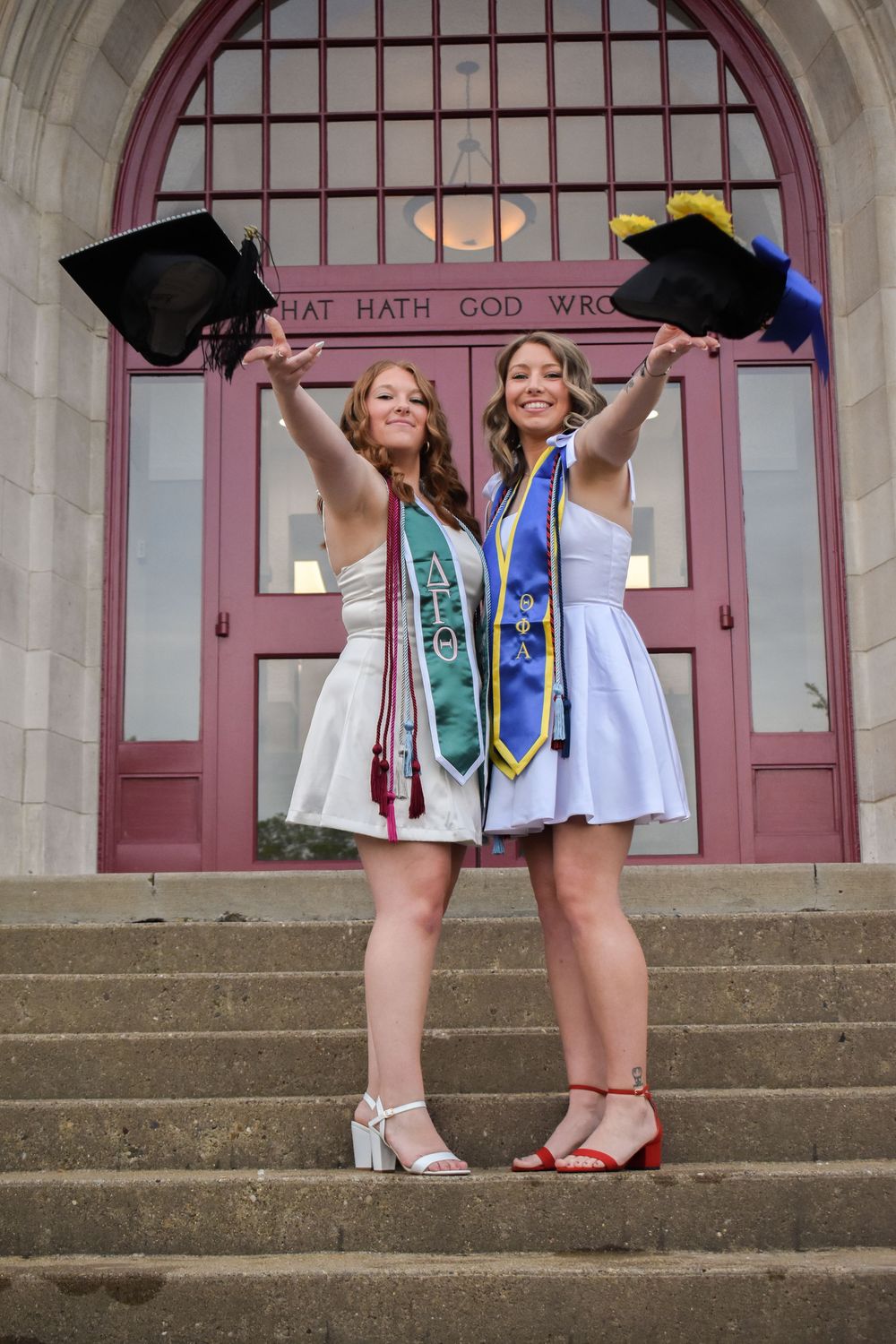 Two smiling female graduates in white dresses hold caps aloft on university steps, wearing colorful honor stoles.