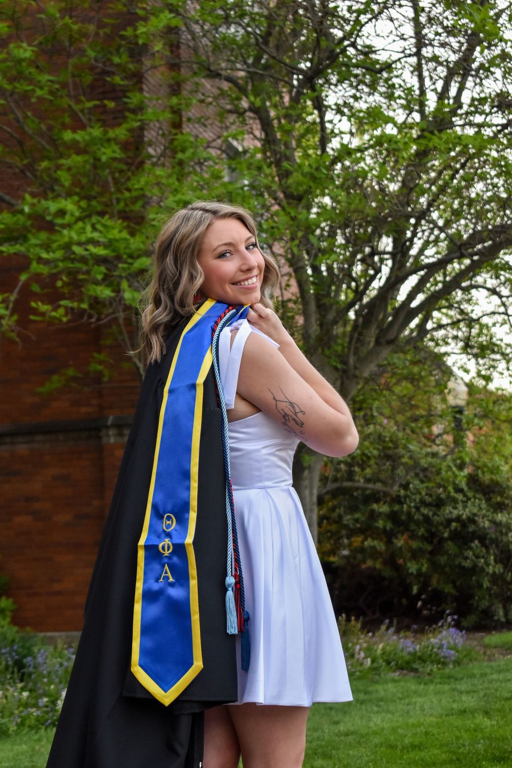 Graduate in white dress and blue academic stole standing outdoors by brick building during commencement celebration.