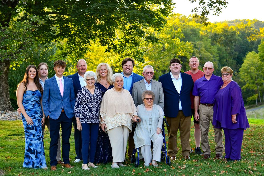 Large family group portrait outdoors on green lawn with trees, dressed in formal attire including blue and purple outfits.