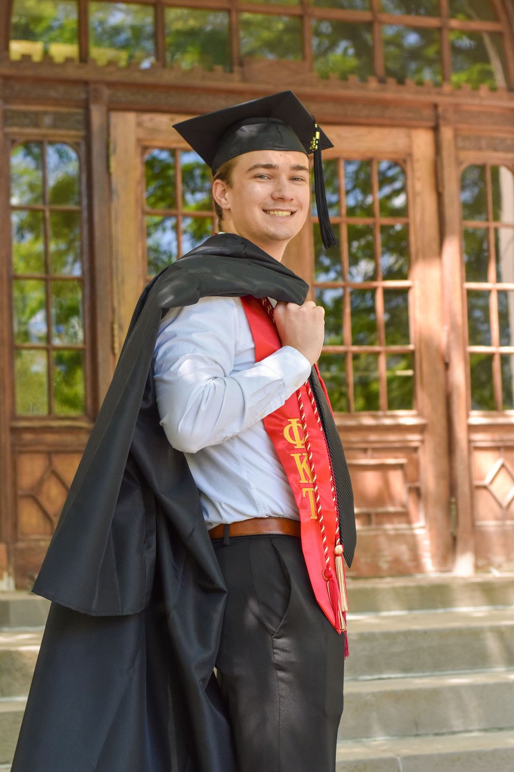 Graduate in cap and gown poses smiling by wooden door wearing red honor stole and black academic regalia.