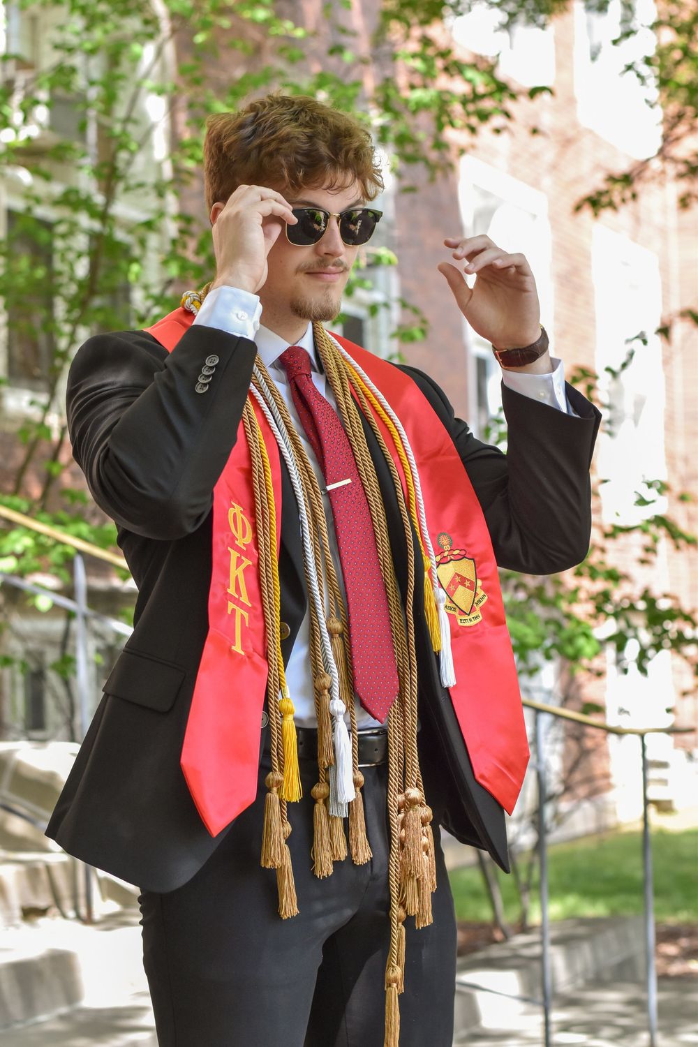 Graduate wearing black suit with red graduation stole and cords poses adjusting sunglasses on sunny spring day.