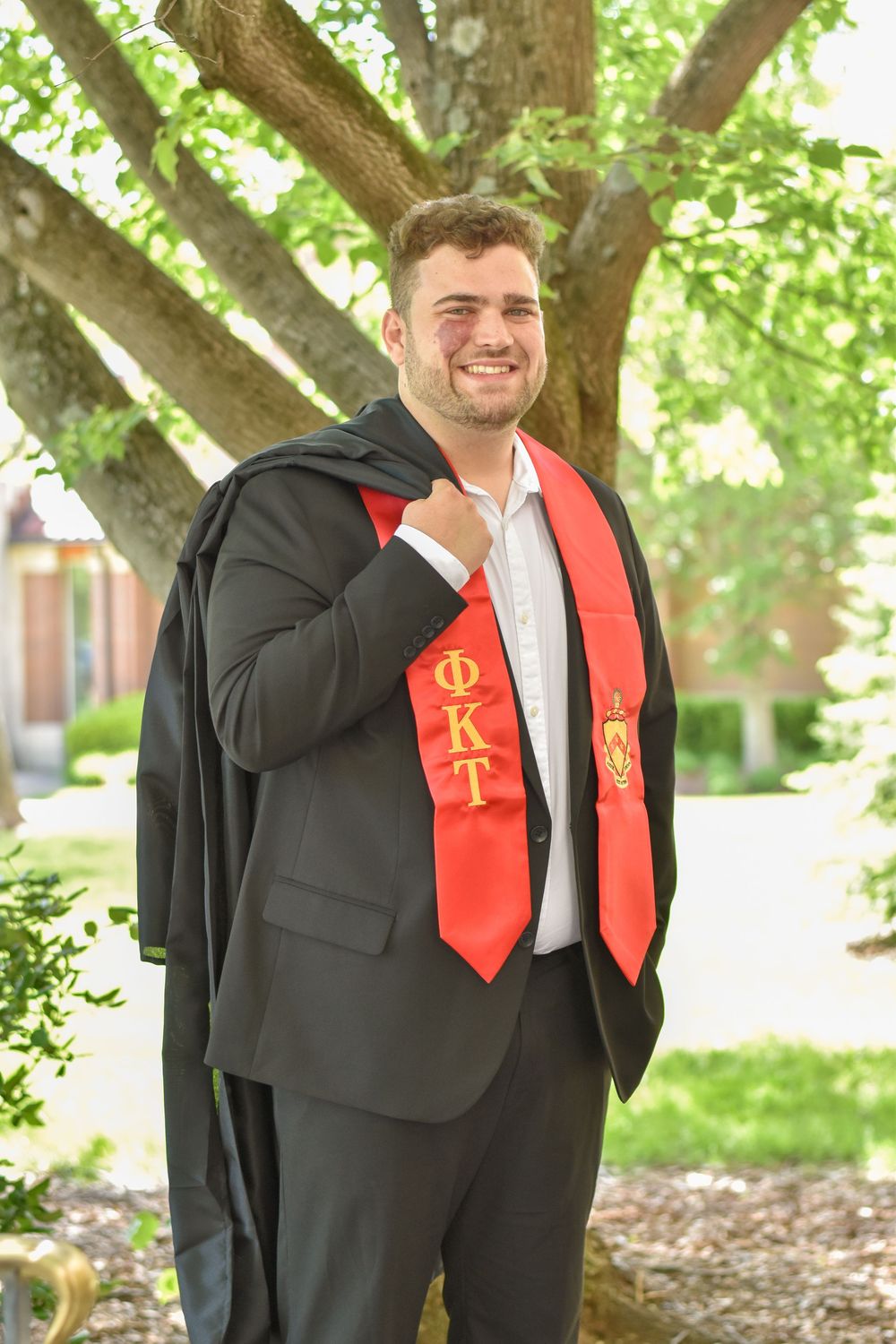 Graduate in black suit with red graduation stole stands smiling under tree branches on sunny day.