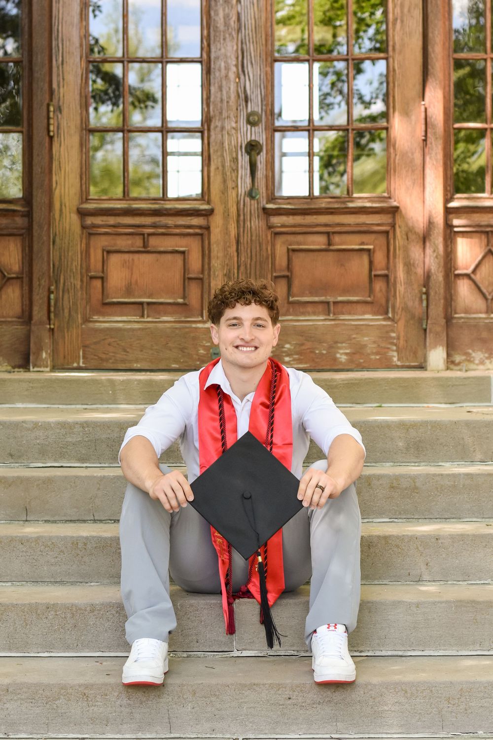 Graduate in white shirt and red stole sits on stone steps holding graduation cap.