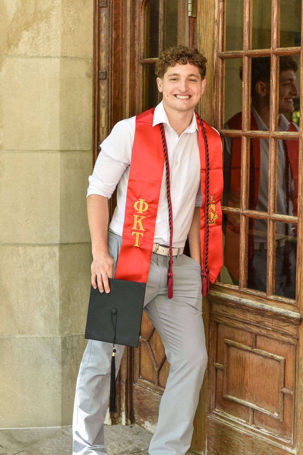 Graduate in red honor stole stands smiling by wooden doorway in formal attire.