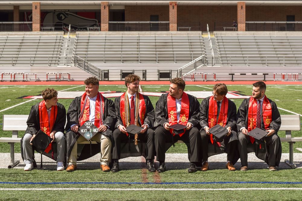 Six graduates wearing red and black stoles sit together on a bench at a stadium football field.