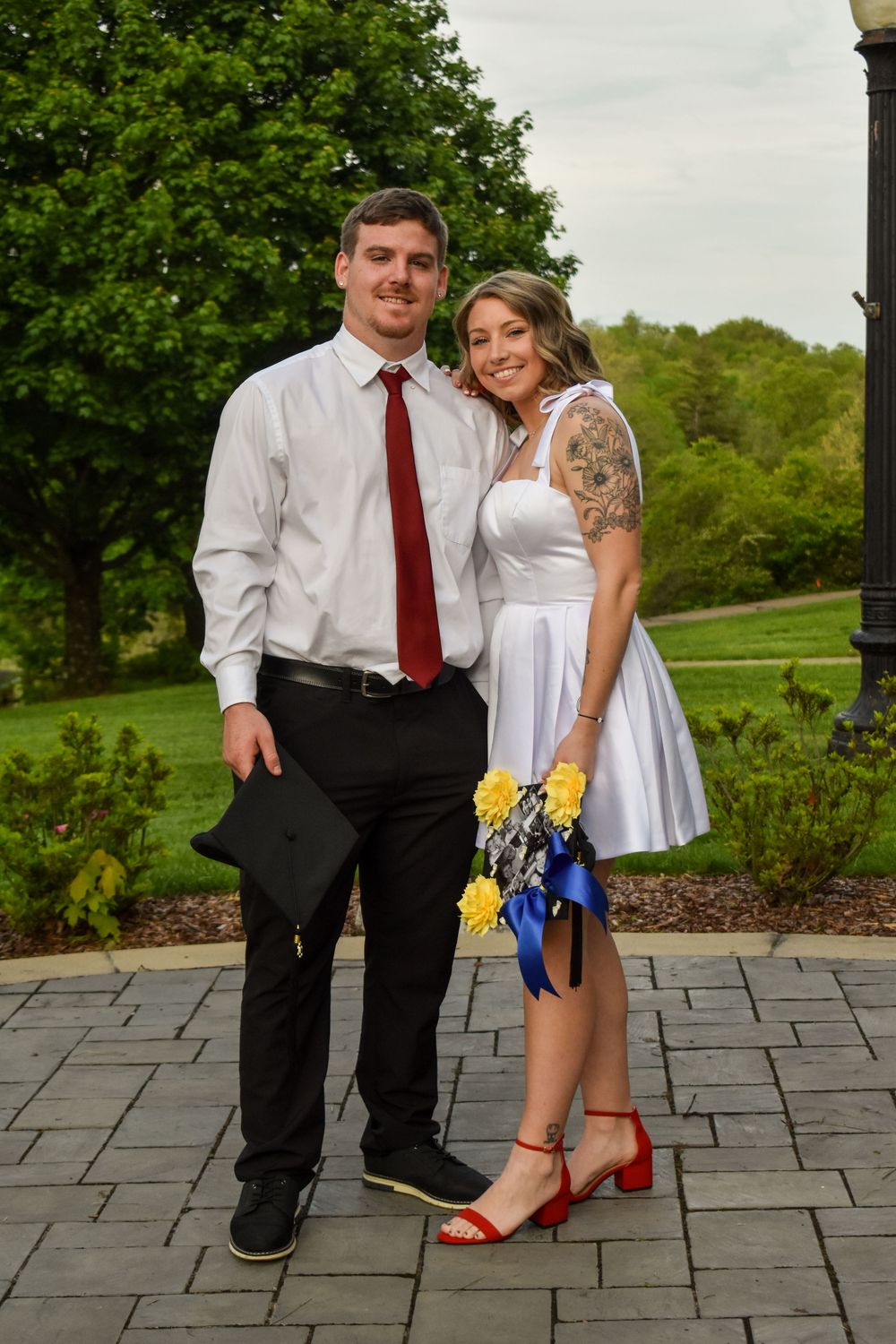 A couple poses together on a stone patio surrounded by greenery, wearing semi-formal attire and holding a blue bouquet.