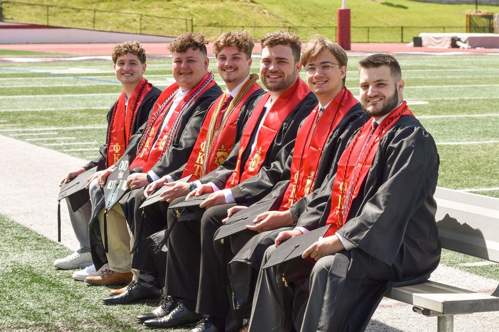 A group of volunteers sit on bleachers wearing red and black jackets on a sports field during a sunny day.
