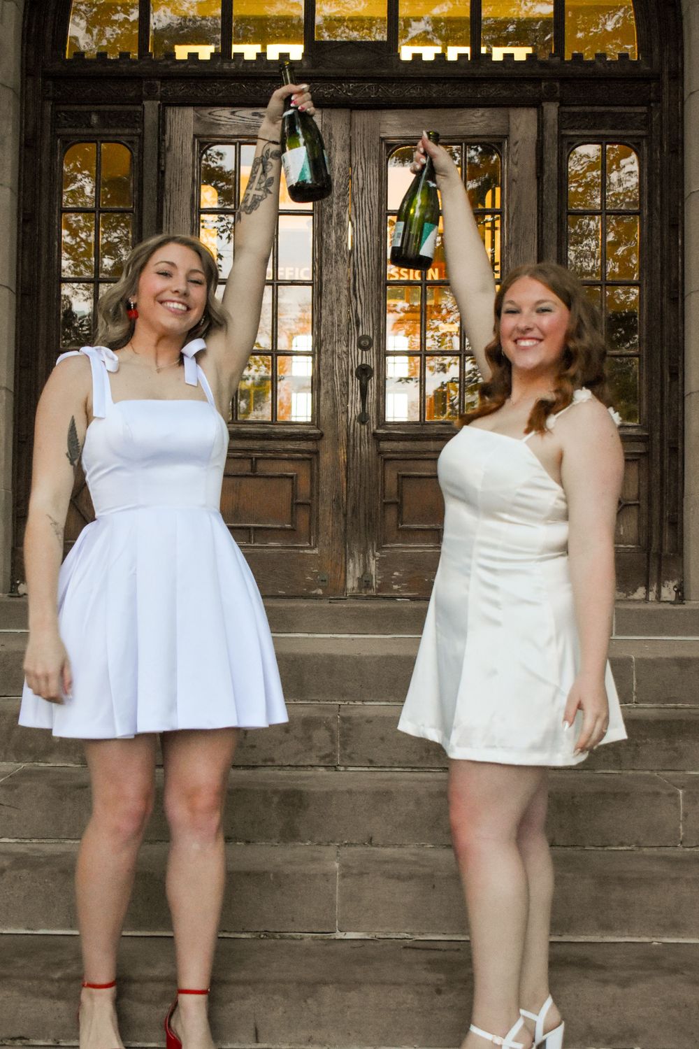 Two friends in white dresses hold up bottles in a celebratory pose in front of vintage wooden doors.