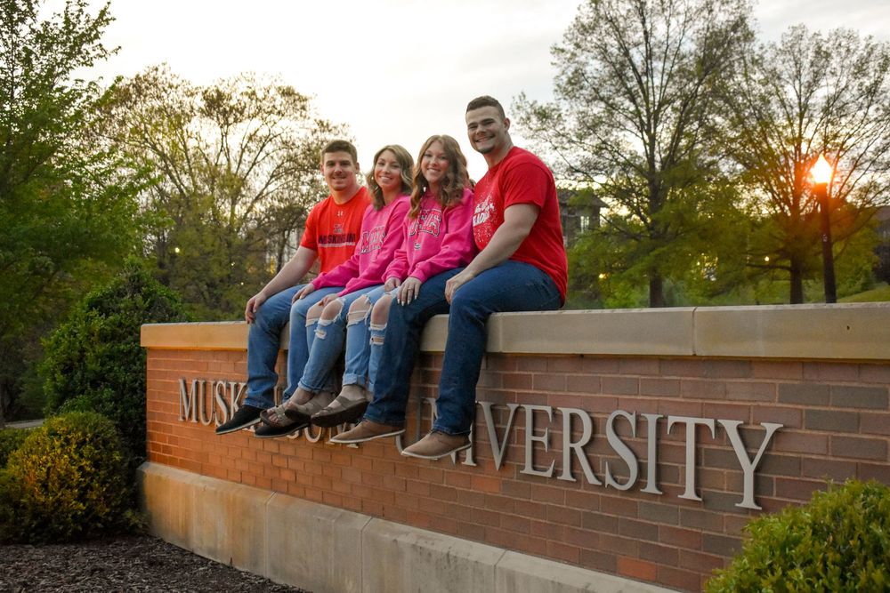 A group sits casually on a brick wall sign with university lettering during sunset outdoors.