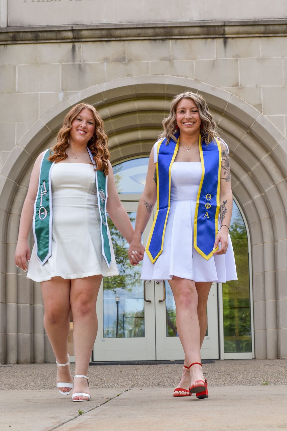 Two graduates in white dresses and graduation stoles hold hands while standing under an arched doorway.
