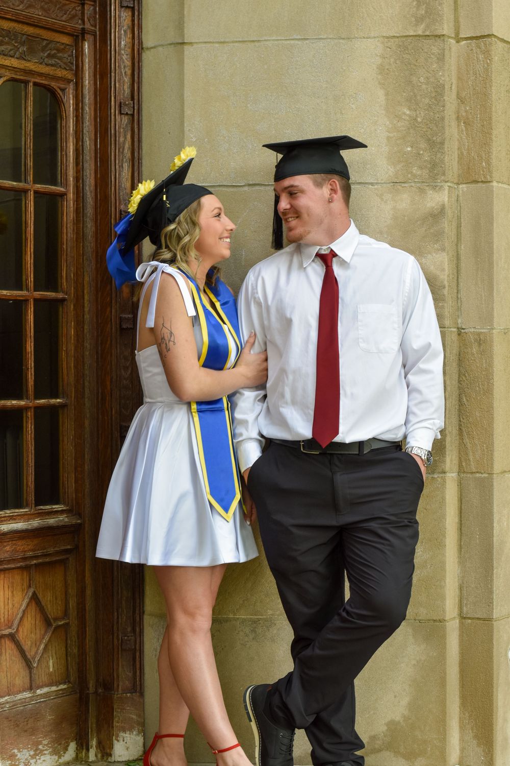 Two graduates wearing caps and gowns share a candid moment together leaning against a stone building.