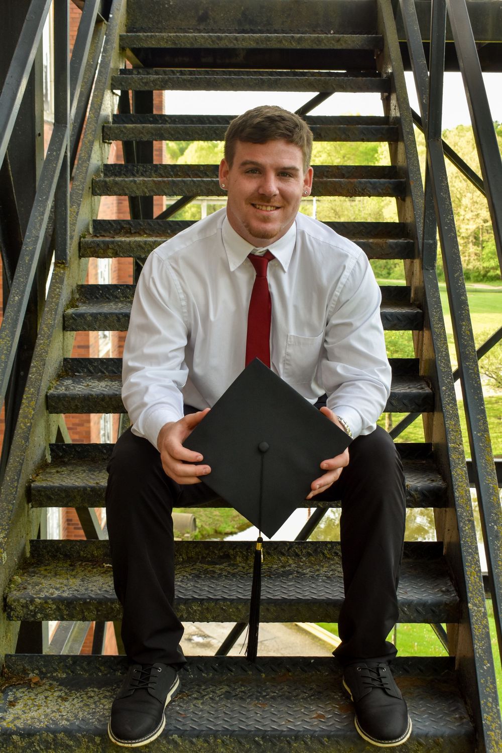 Graduate in formal attire sits on metal stairs holding mortarboard cap during outdoor graduation photoshoot.