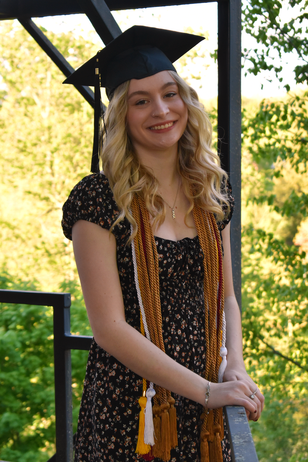 Graduate wearing cap and floral dress with gold honors cords standing in outdoor gazebo.