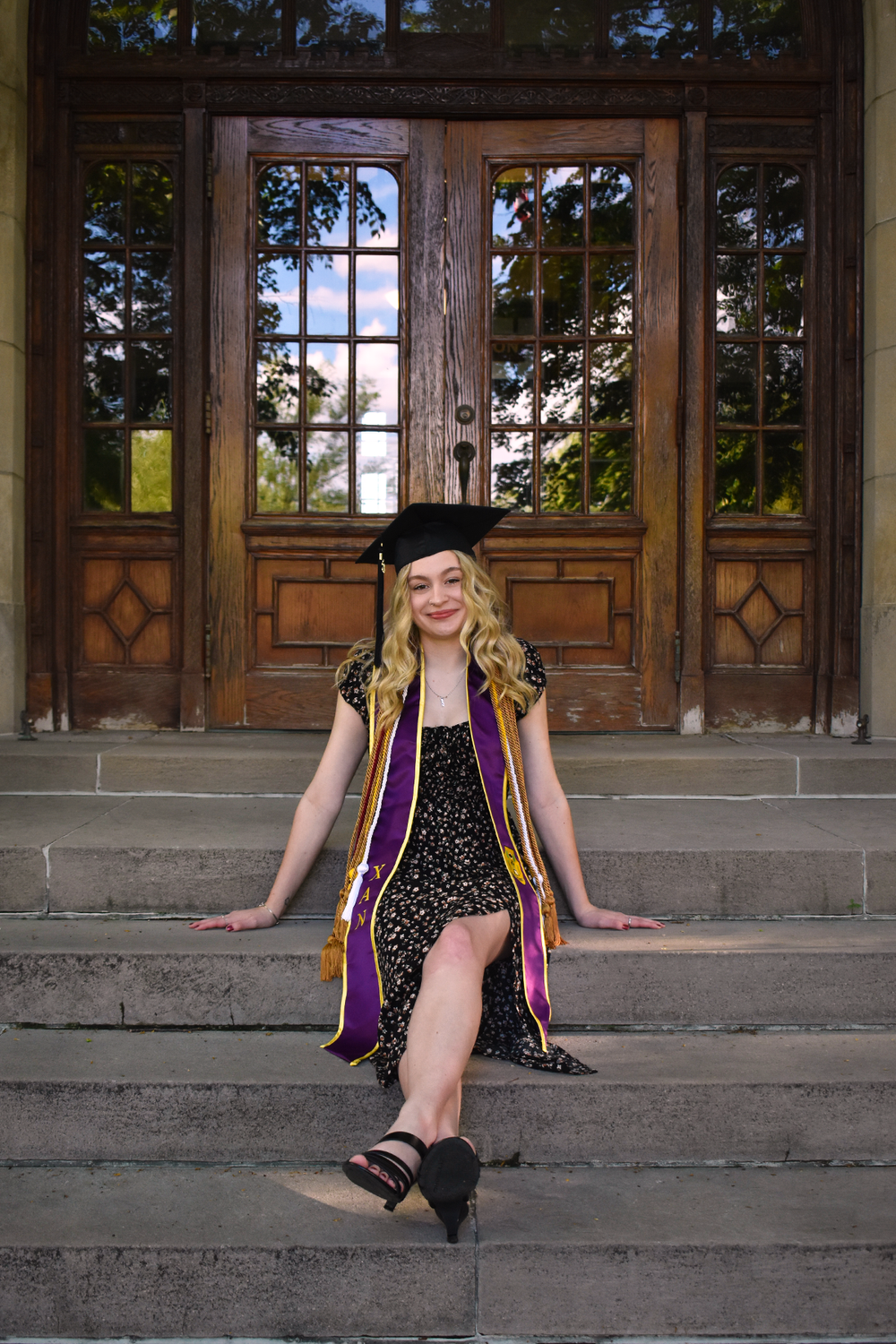 Graduate in black dress and academic regalia sitting on historic building steps.